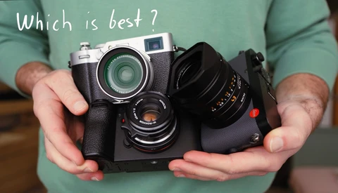 Hands holding a rangefinder camera with interchangeable lens and accessories, posed against a green shirt.