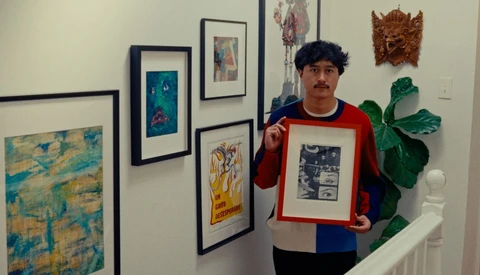 Artist holding a framed black and white print in a gallery space surrounded by various artworks.