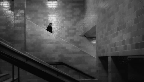 Solitary figure in dark clothing walking down a steep concrete staircase in a monochromatic architectural space.