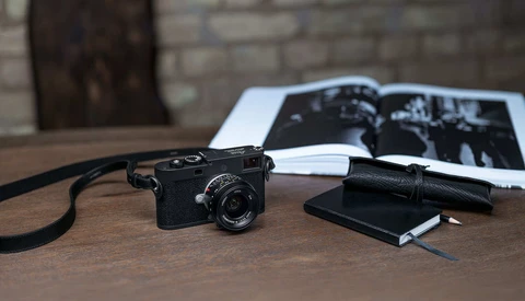 Black rangefinder camera with strap on wooden desk beside photographs and notebook