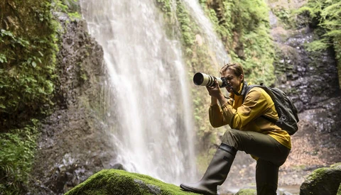 Photographer in yellow jacket using telephoto lens at misty waterfall with moss-covered rocks