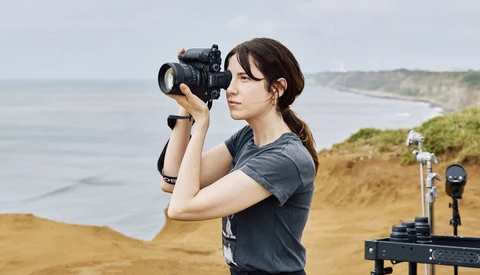 Female photographer holding DSLR camera to her face while standing on coastal cliff overlooking ocean
