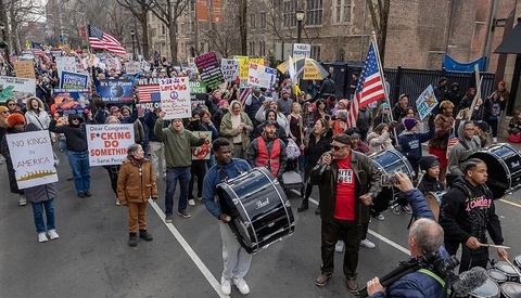 Large crowd of protesters marching down an urban street, carrying signs and American flags.