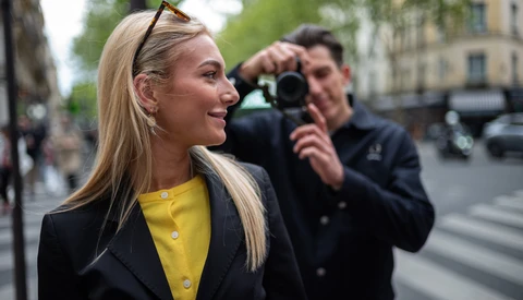 Woman in yellow shirt and black blazer smiling while photographer captures her portrait on an urban street.