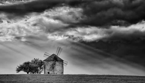 A solitary windmill stands in an open field beneath a dramatic cloudy sky in black and white.