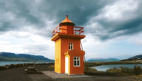 Bright orange and yellow lighthouse structure standing alone on dark sand with mountains and water visible in the background under stormy skies.