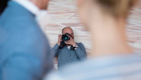 Photographer holding a camera and shooting while two people pose in the foreground, with a brick wall background.