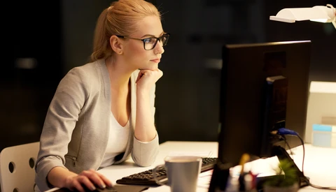 Woman with glasses working at a desk in front of a computer monitor at night.