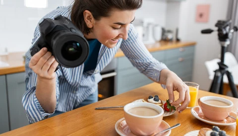 Food photographer holding DSLR camera while composing flat lay of coffee cups and pastries on wooden table