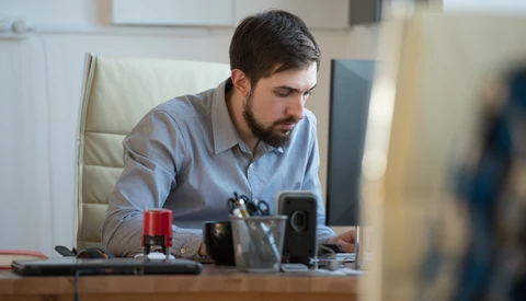 Man in light blue shirt seated at desk, looking down at work in modern office setting.