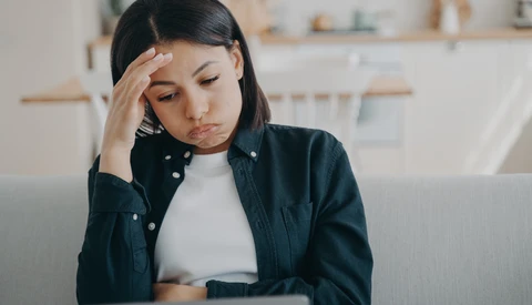 Woman with dark hair wearing a dark shirt holds her head while looking down at a laptop with a concerned expression.