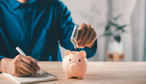 Person in blue shirt dropping a coin into a pink piggy bank while writing in a notebook.