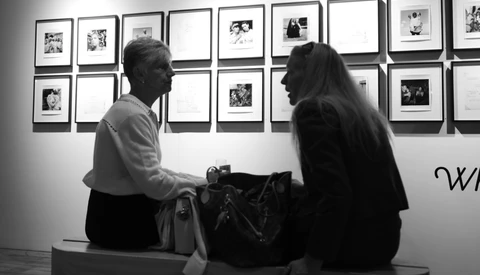 Two people seated on a bench viewing framed photographs displayed on a gallery wall in black and white.