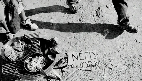 Overhead shot of person sitting on pavement with food and sign reading "NEED WORK"