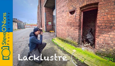 Photographer crouching to compose a shot of a moss-covered ground strip against weathered brick industrial building.