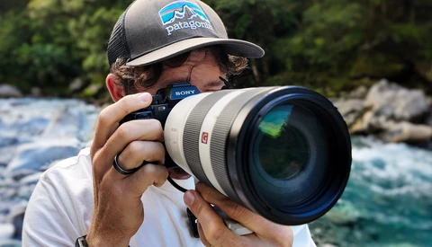 Photographer holding Sony camera with telephoto lens near flowing turquoise water and rocky shoreline.