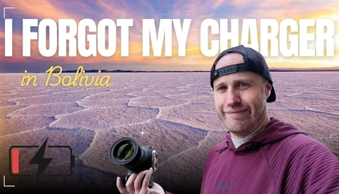 Photographer holding a camera with a depleted battery indicator, standing in front of a vast salt flat at sunset.