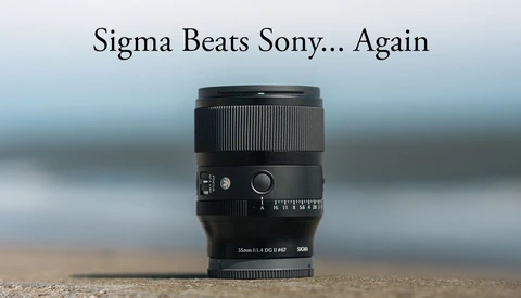 Sigma 35mm f/1.4 DG HSM lens positioned upright on sand against a blurred coastal background.