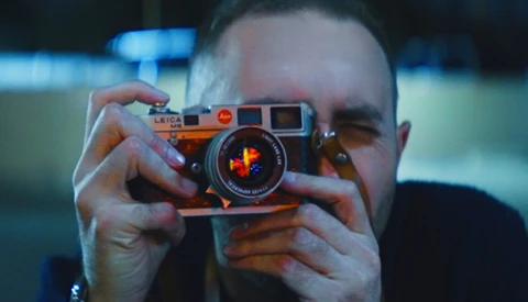Photographer holding an orange rangefinder camera up to their face, obscuring their head while composing a shot.