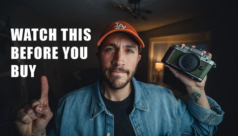 Man wearing LA cap holding a rangefinder camera while pointing at the viewer indoors.
