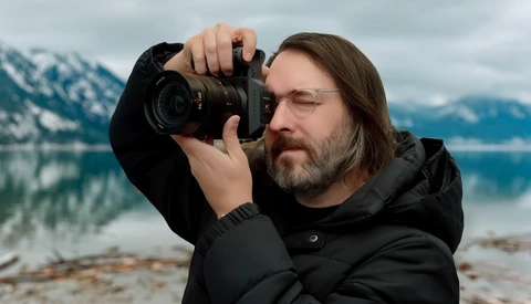 Photographer holding a DSLR camera to his eye while composing a shot of a mountain lake landscape.