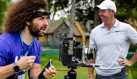 Filmmaker with camera on tripod discussing with golfer on outdoor course.
