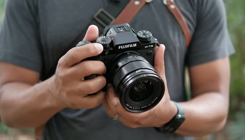 Photographer holding a Fujifilm X-T5 camera with attached lens, wearing a gray shirt and brown camera strap.