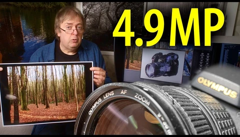 Photographer holding printed forest landscape photo next to Olympus lens and camera equipment on desk.