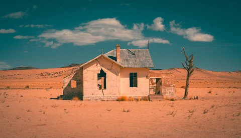 Abandoned house with peeling pink walls and dark windows stands alone in vast, arid desert landscape.