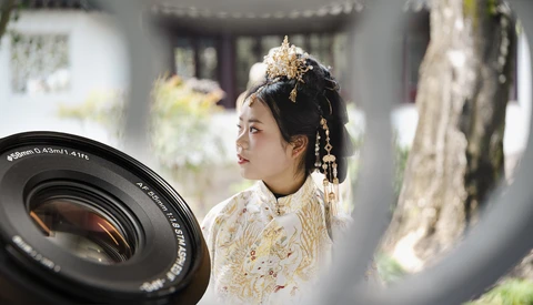 Woman in ornate white and gold traditional dress with elaborate gold headdress, photographed through a camera lens in soft natural light.