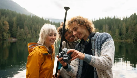 Three people holding a paddle while standing in a boat on a calm alpine lake surrounded by forested mountains