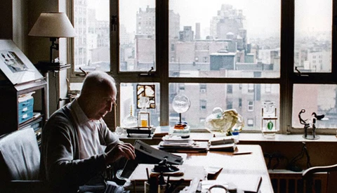 Man working at typewriter at desk overlooking urban skyline through large windows