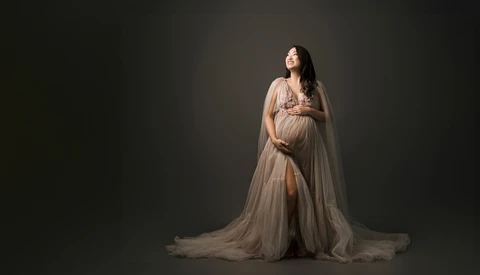 Pregnant woman in flowing sheer cape gown gazing upward against dark background