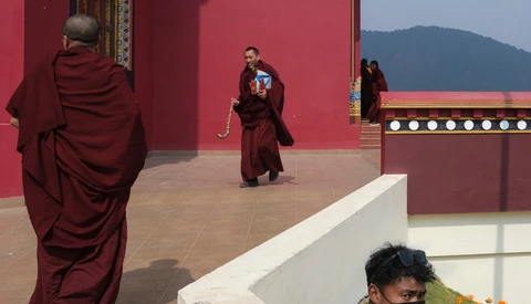 Buddhist monks in maroon robes walking across a courtyard with deep red walls and mountain backdrop