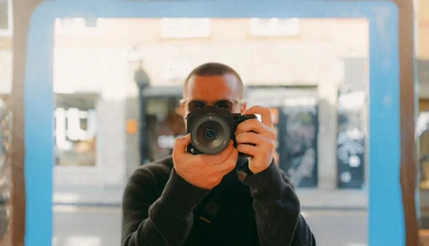 Photographer holding a mirrorless camera up to their face, viewed through a blue-framed mirror or reflective surface.