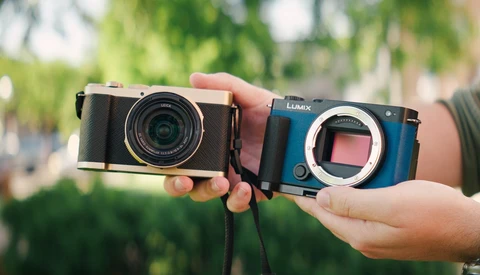 Hands holding a black rangefinder camera and blue Lumix mirrorless camera body side by side outdoors