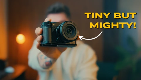 Photographer holding a compact mirrorless camera toward the viewer with overlaid text reading 'TINY BUT MIGHTY!'