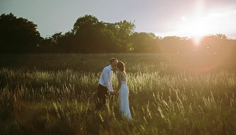 Couple kissing in golden hour sunlight amid tall grass field with trees silhouetted in background
