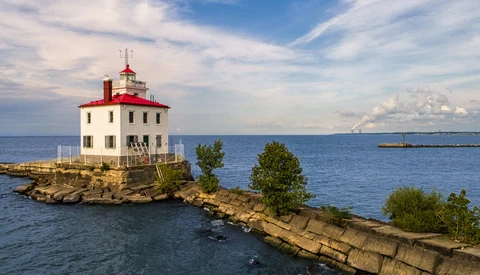 Red and white lighthouse on rocky breakwater surrounded by blue water under partly cloudy sky