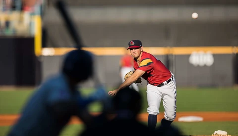 Baseball pitcher in red uniform mid-delivery with batter in foreground out of focus