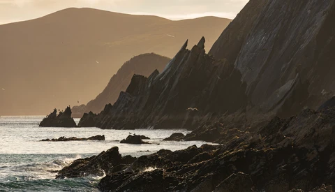 Dramatic coastal cliffs with castle ruins silhouetted against misty mountains and golden hour light