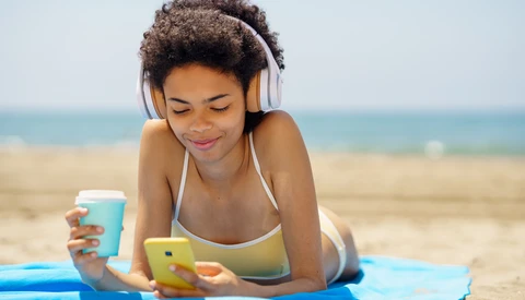 Woman wearing headphones lying on beach towel holding smartphone and beverage