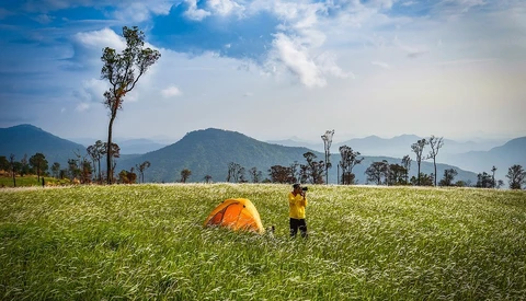 Hiker in yellow jacket beside orange tent in expansive green field with mountains and palm trees