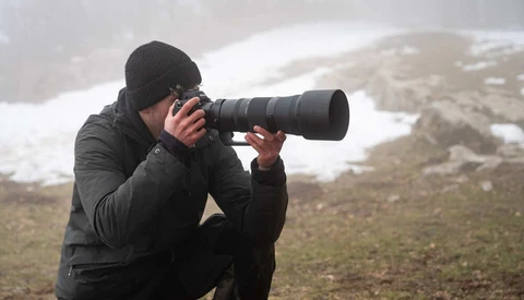 Photographer in black winter gear crouching while composing shot with telephoto lens in snowy mountain terrain