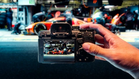 Hand holding mirrorless camera displaying racing pit crew on LCD screen with blurred race car in background