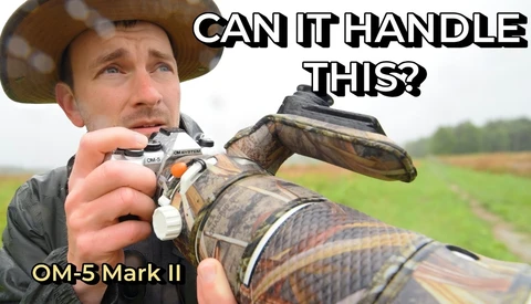Photographer in cowboy hat holding OM-5 Mark II camera with large telephoto lens in muddy field
