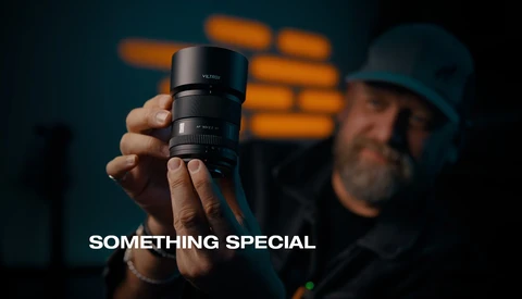 Photographer holding a telephoto lens toward camera with brick wall backdrop