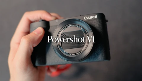 Hand holding Canon PowerShot V1 compact camera with zoom lens against gray background