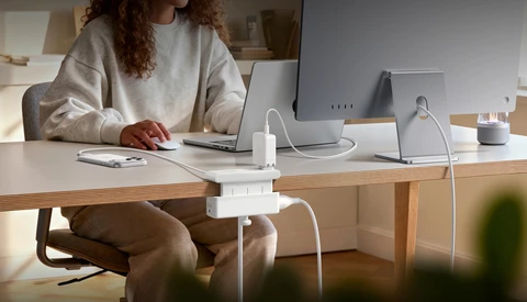 Woman at wooden desk working on laptop surrounded by white tech accessories and charging devices