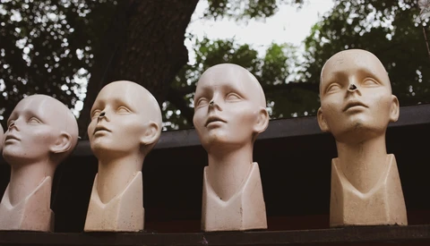 Four pale sculptural heads displayed on a dark shelf against a natural backdrop